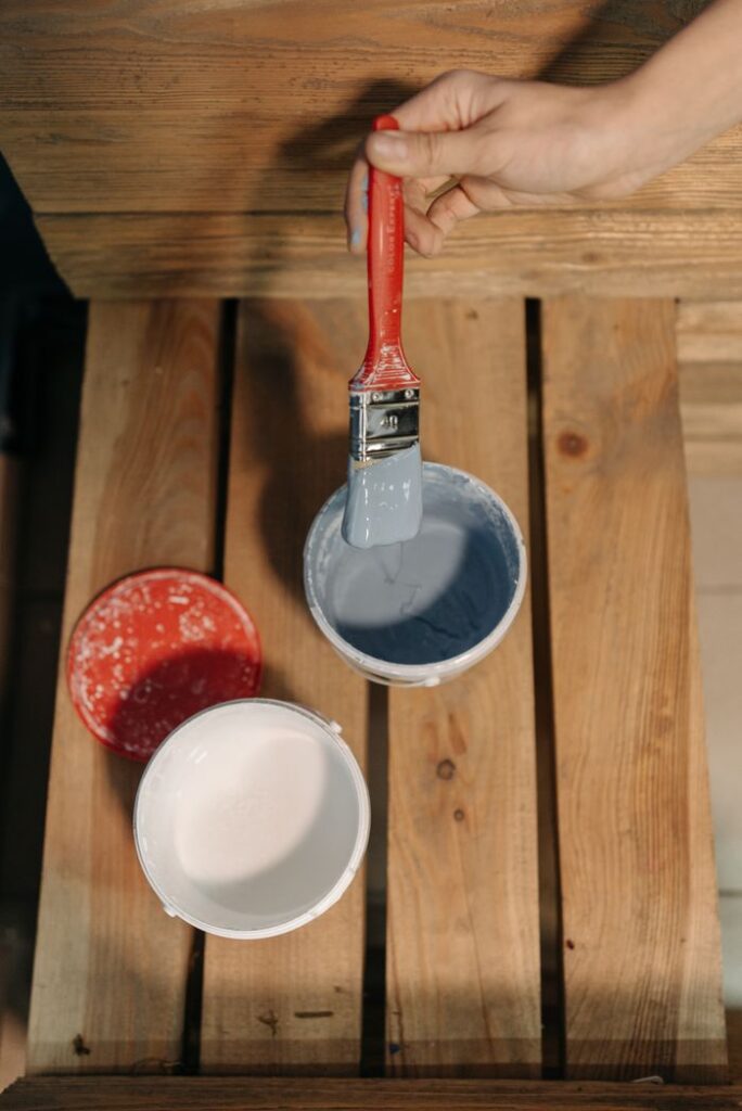 A hand holds a paintbrush over open paint cans on a wooden table. Artistic setup perfect for DIY home projects.